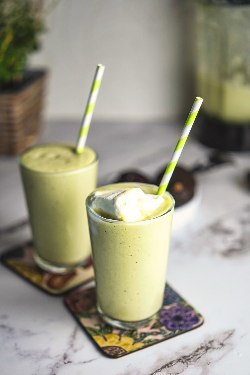 Two glasses of creamy avocado milkshake with striped straws, topped with whipped cream, on a marble countertop.