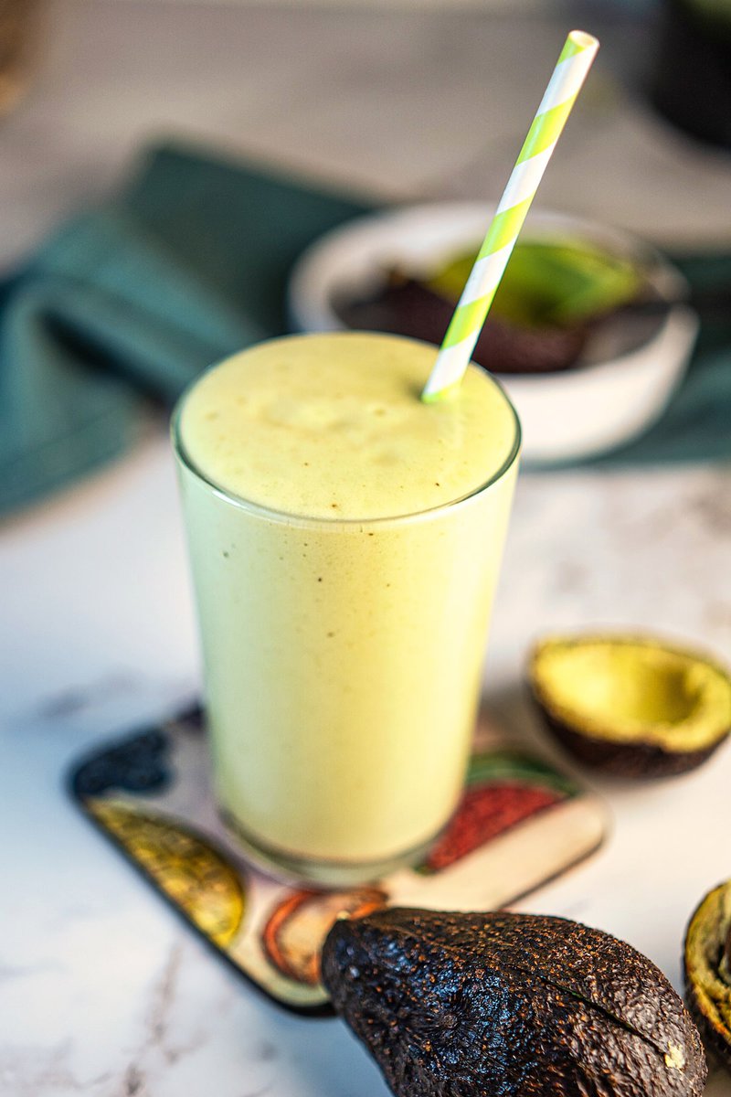 A creamy avocado milkshake in a glass with a striped straw, surrounded by avocado halves.