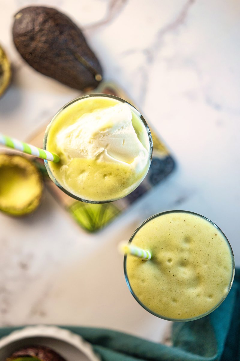 Two glasses of creamy avocado milkshake with striped straws, surrounded by ripe avocados on a marble surface.