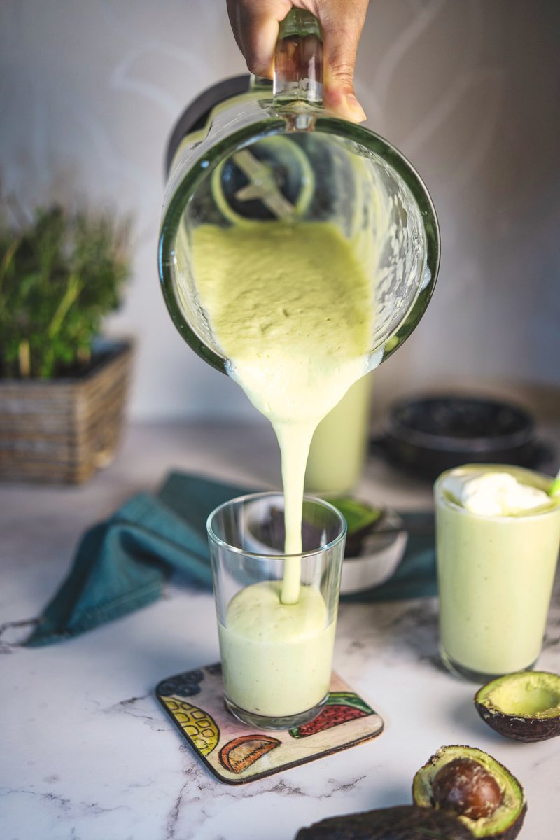 A creamy avocado milkshake being poured into a glass, surrounded by fresh ingredients.