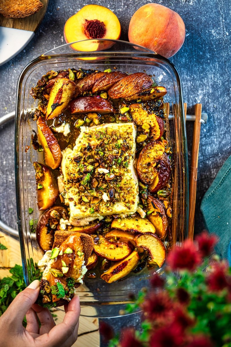 Baked peach feta being served with toasted bread, creamy feta, juicy caramelized peaches, roasted pistachios, fresh mint, and honey