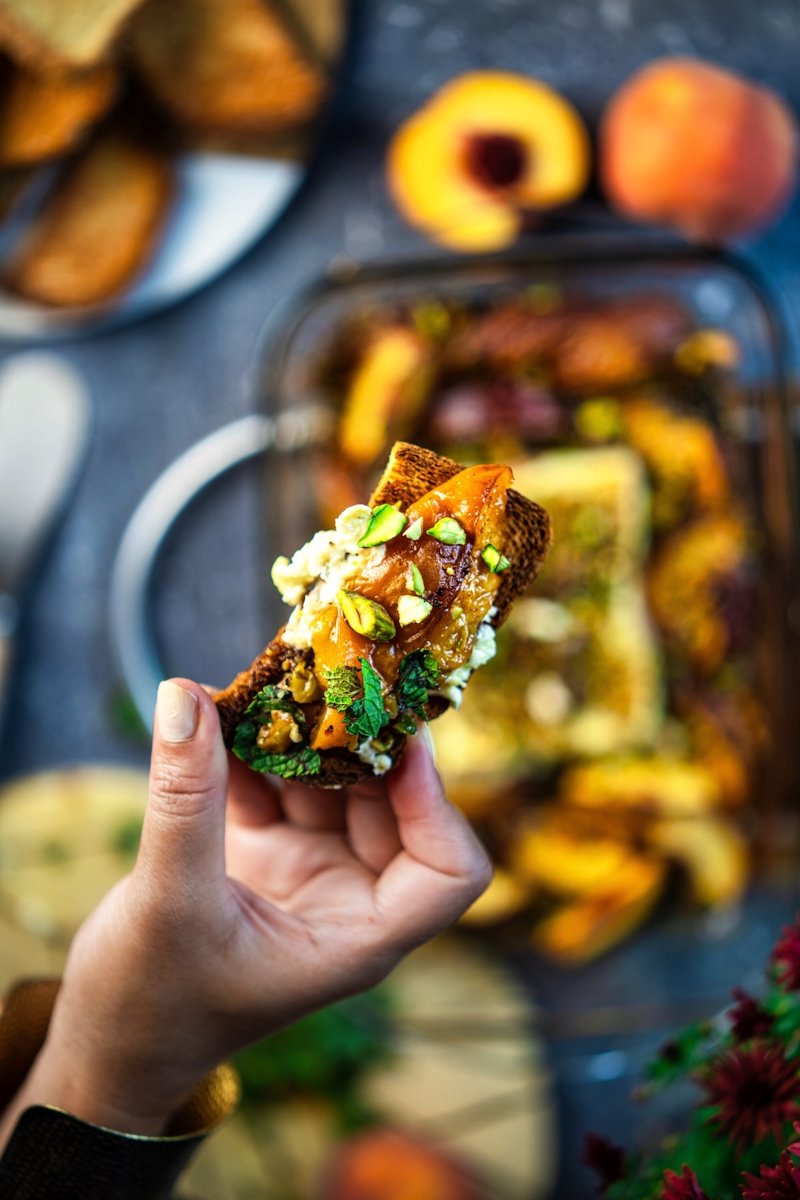Close-up of toasted bread topped with creamy baked feta, caramelized peach, roasted pistachios, fresh mint, and honey