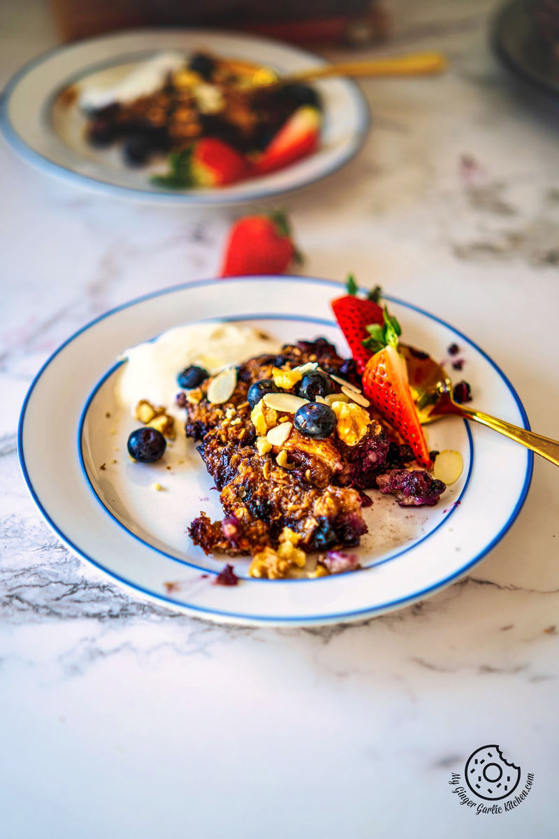 a white plate with a slice of banana blueberry baked oats, walnuts, almonds, yogurt, strawberries and blueberries