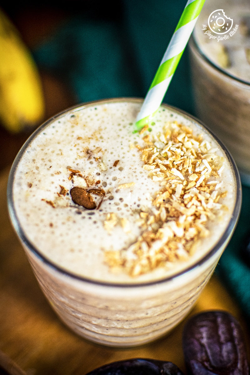 A frothy banana date smoothie topped with granola, cinnamon, and a green-striped straw in a clear glass.