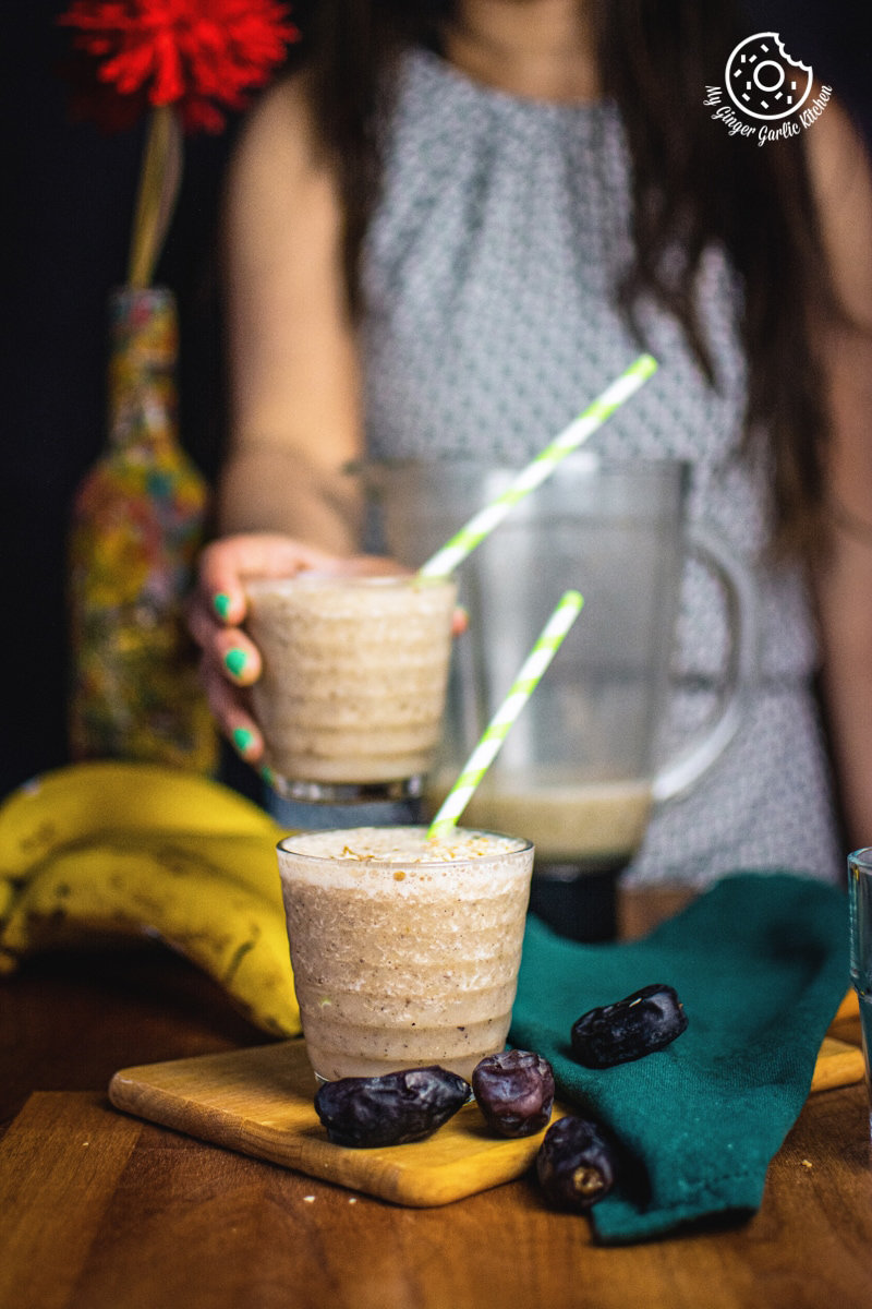 Hand holding a glass of banana date smoothie, with a pitcher and a bowl of fresh bananas and dates in the background.