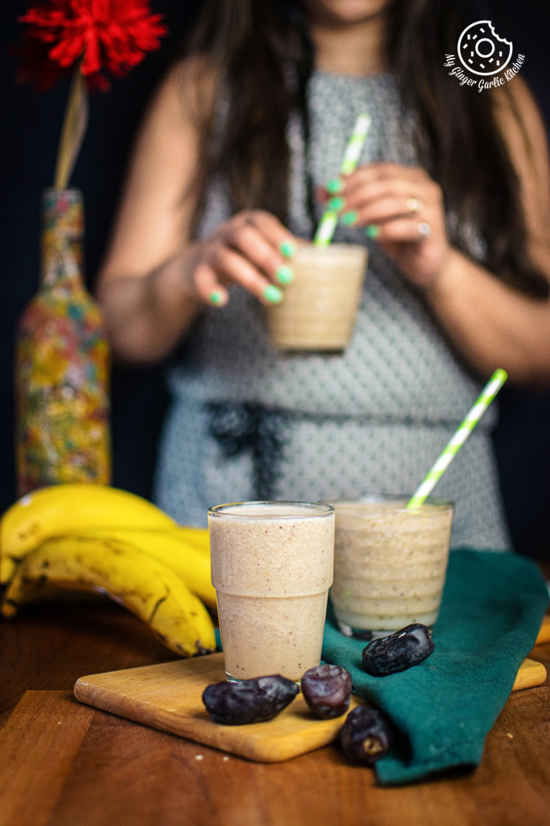 A female holding banana date smoothie glass, with fresh bananas and dates laid out on a wooden board.