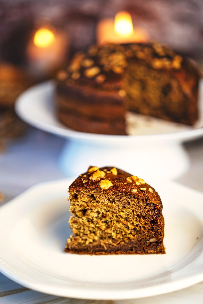 A close-up of a slice of eggless banana date walnut cake on a white dish, with the whole cake and candles in the blurry background.