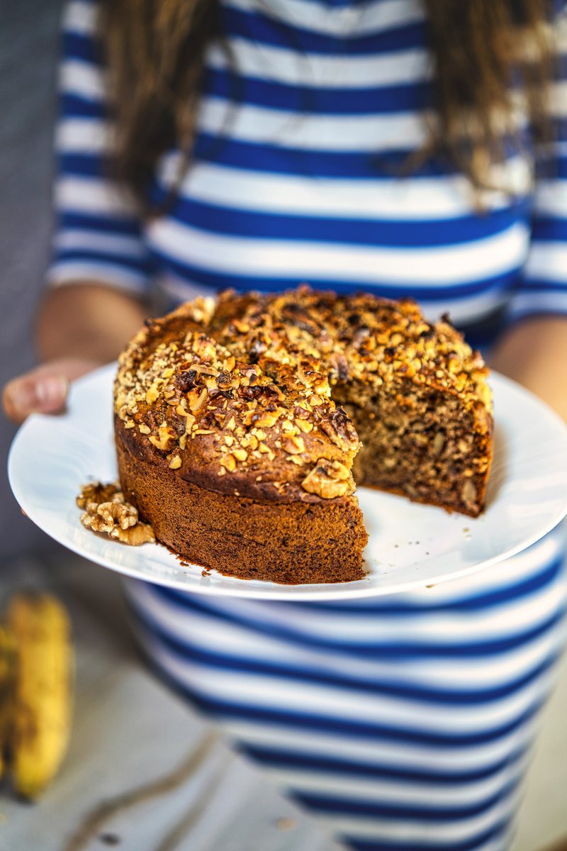 A person holding a plate with a sliced eggless banana date walnut cake, garnished with crushed walnuts.