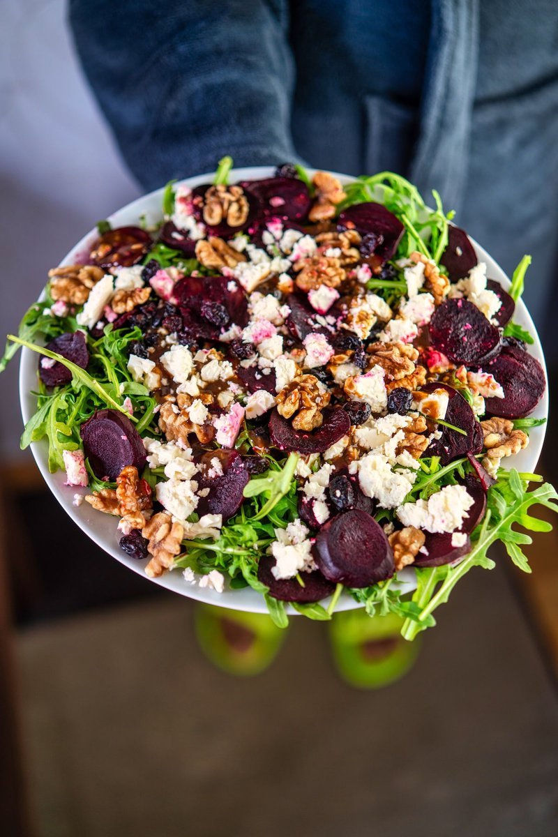 Person holding a colorful beetroot salad with crumbled goat cheese, walnuts, and arugula on a white plate.