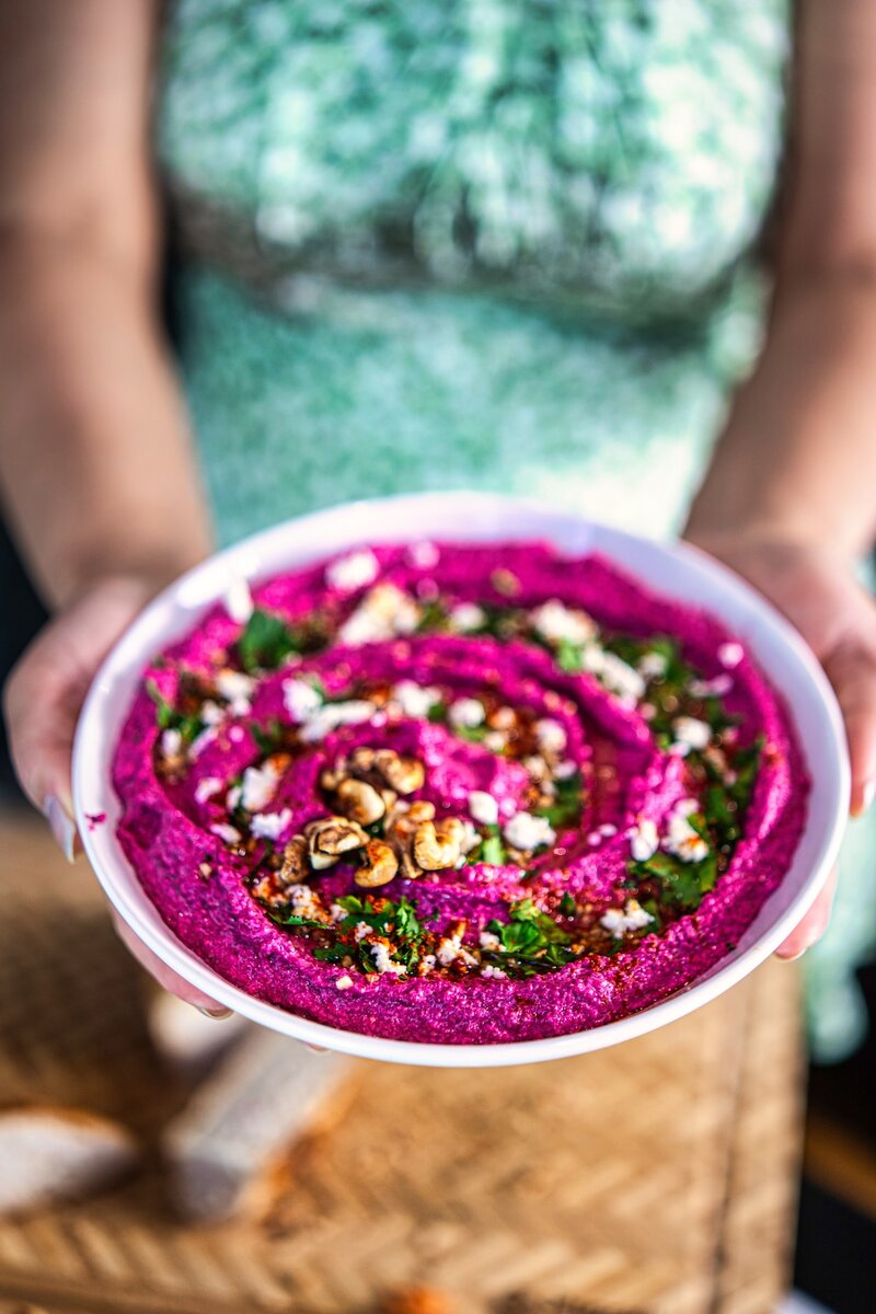 Creamy beetroot walnut feta dip in a bowl held by hands, topped with herbs, feta, and olive oil