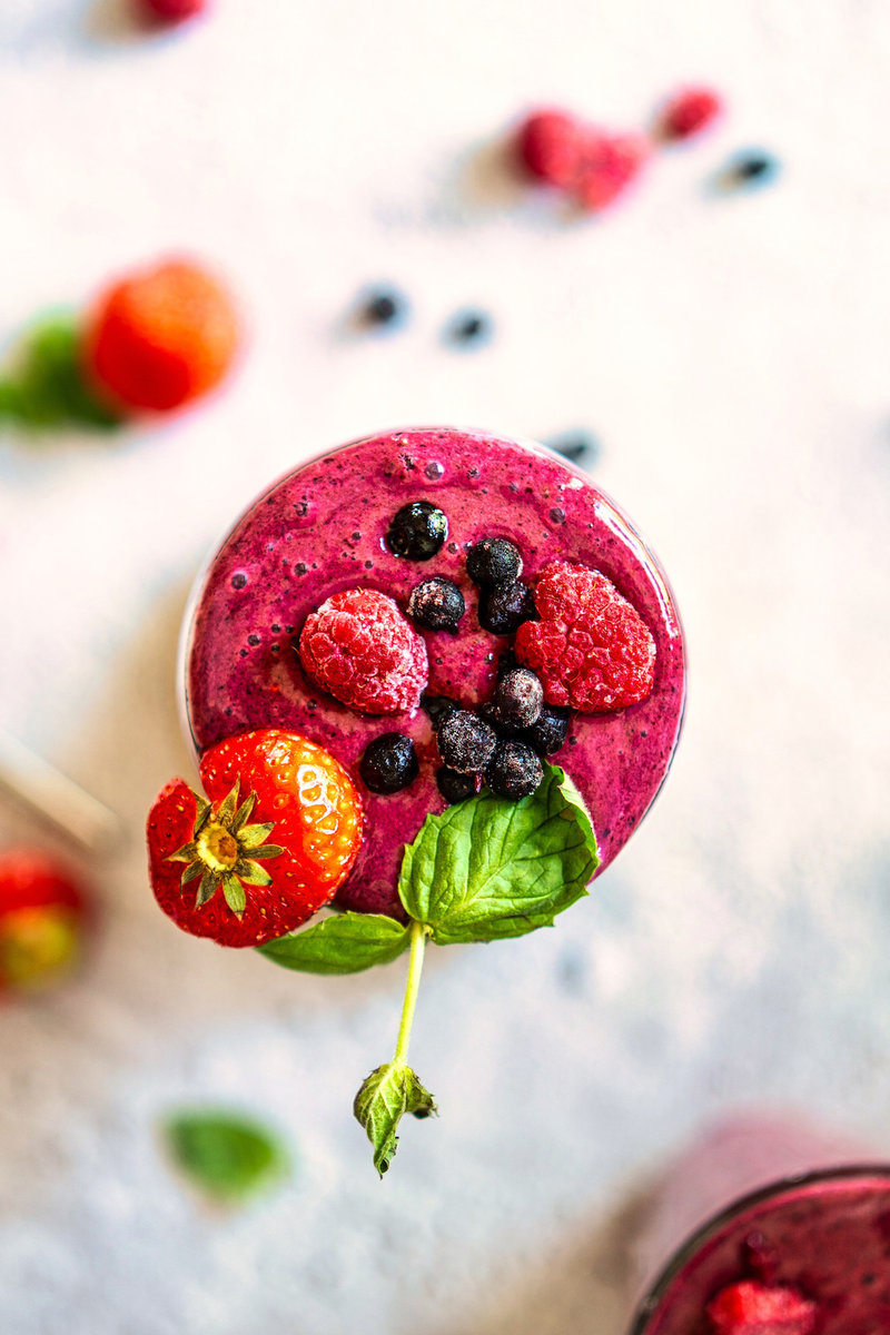 Top view of a berry smoothie garnished with fresh strawberries and blueberries, with additional fruits scattered around.