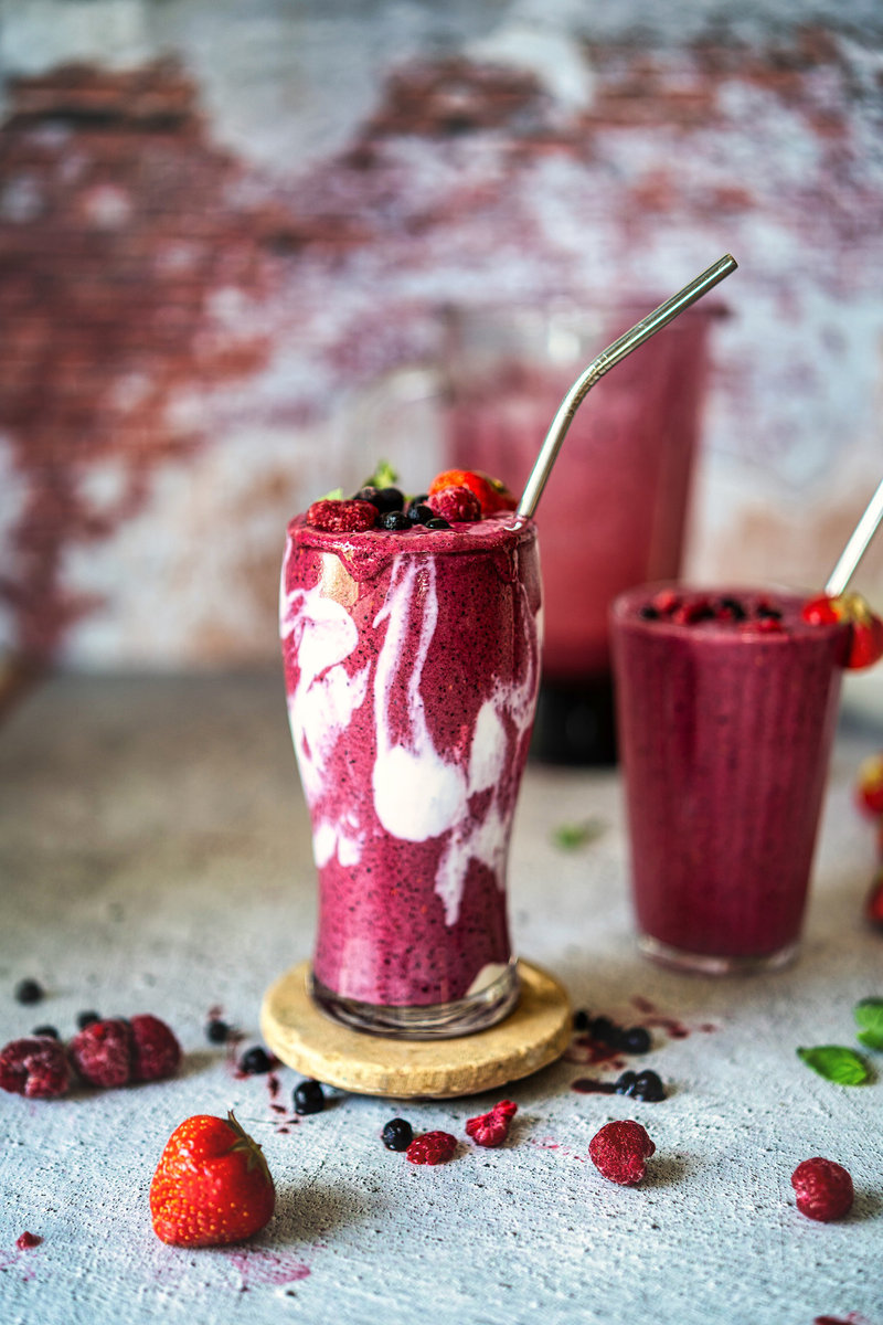 Vibrant mixed berry smoothie served in a tall glass with a metal straw, surrounded by scattered berries on a rustic surface.