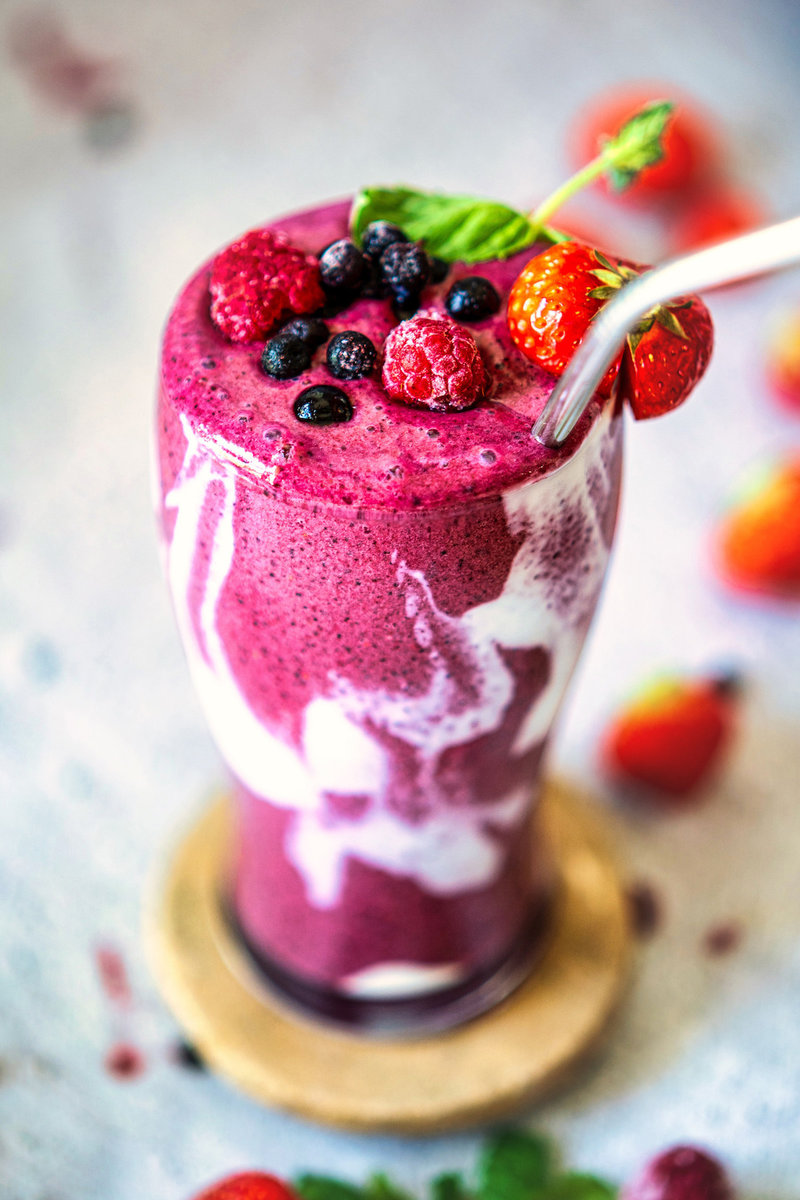 Close-up of a berry smoothie in a glass, topped with strawberries, raspberries, and blueberries, with a metal straw.