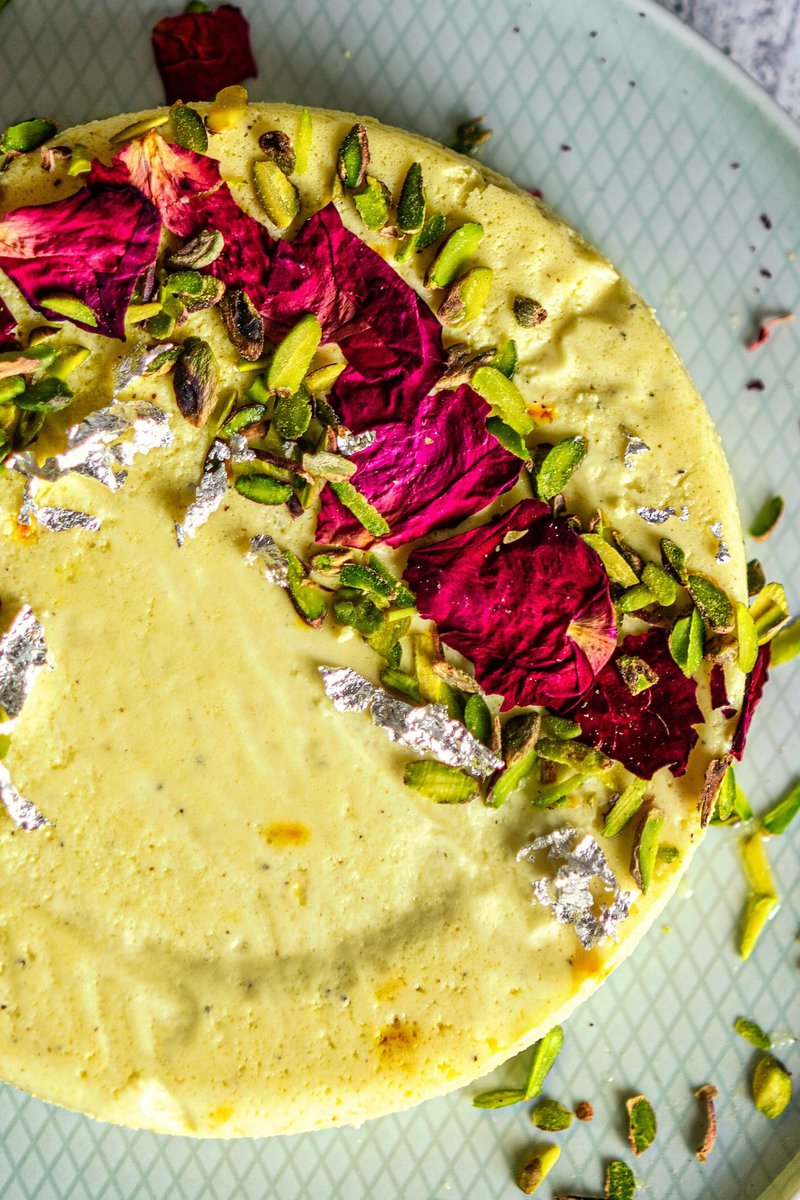 Close-up overhead view of bhapa doi with one slice taken out, garnished with pistachios and rose petals on a pale patterned plate.