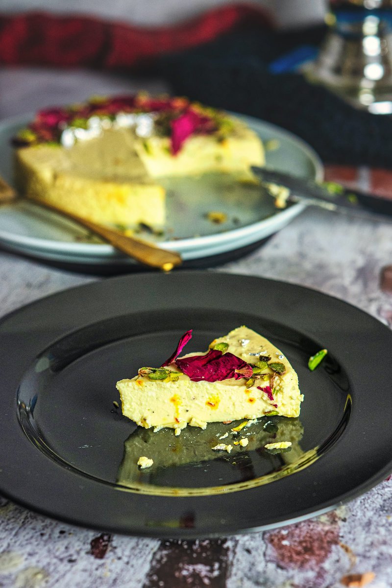 A slice of bhapa doi garnished with pistachios and rose petals on a black plate with the remaining portion in the background. Bhapa Doi is typically made by steaming a mixture of yogurt, milk, sugar, and spices in a mold. The dessert is then chilled and served.