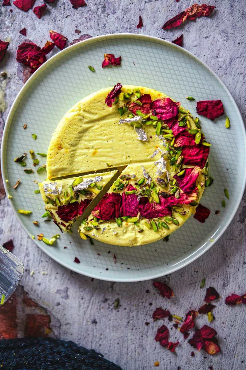 Overhead view of a bhapa doi with a slice removed, showing pistachios and rose petals garnish on a pale patterned plate. This picture is of a popular Indian dessert called Bhapa Doi. It is a creamy, yogurt-based dessert that is often flavored with cardamom and saffron.