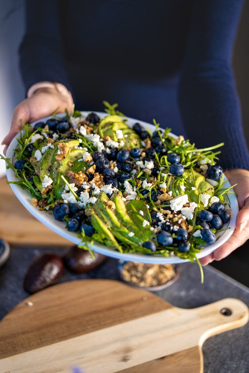 Hands holding a bowl of blueberry avocado salad with arugula, walnuts, and lemon dressing