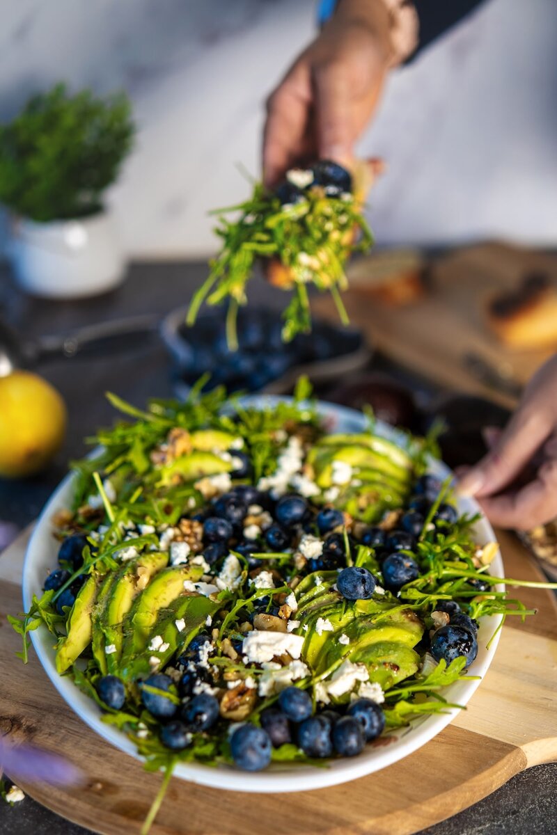 Fresh blueberry avocado salad being served with arugula, walnuts, and vegan feta on a wooden board