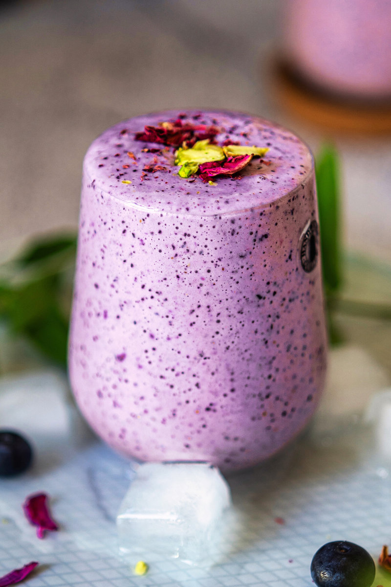 A close-up of a chilled blueberry lassi garnished with rose petals and pistachios, with ice cubes around the base.