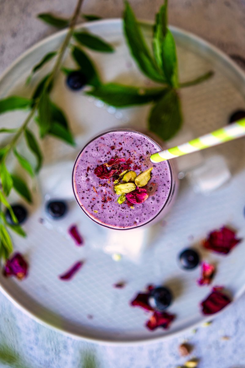 Top view of a blueberry lassi with a straw, garnished with rose petals and pistachios, surrounded by fresh blueberries and green leaves.