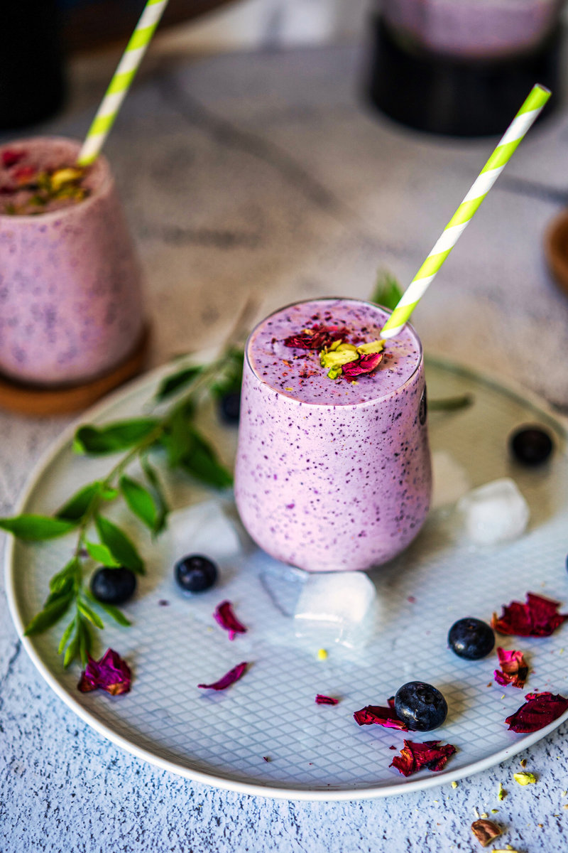 A blueberry lassi with a straw, placed on a tray with fresh blueberries, ice cubes, and rose petals scattered around.