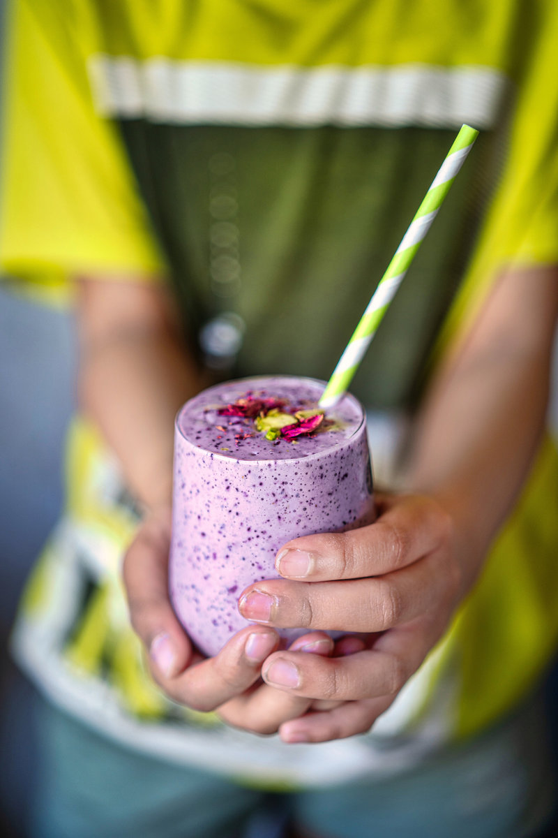 A person holding a blueberry lassi garnished with rose petals and pistachios, with a striped straw in the drink.