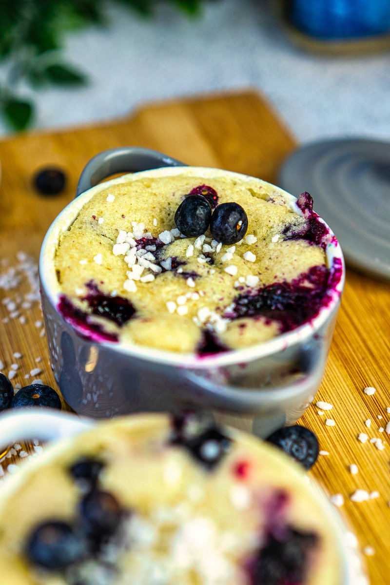 A close-up image of a blueberry mug cake in a gray mug. The cake is topped with blueberries and sugar crystals.