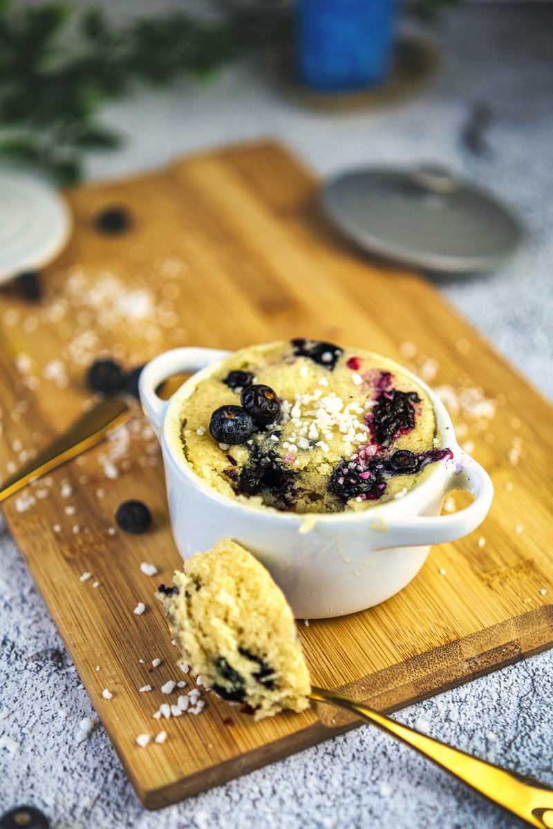 A close-up image of a blueberry mug cake in a gray mug on a wooden table. The cake is topped with fresh blueberries and sugar crystals.