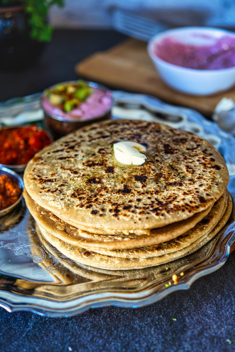 Stack of broccoli parathas topped with a dollop of butter on a silver tray with side dishes in the background.