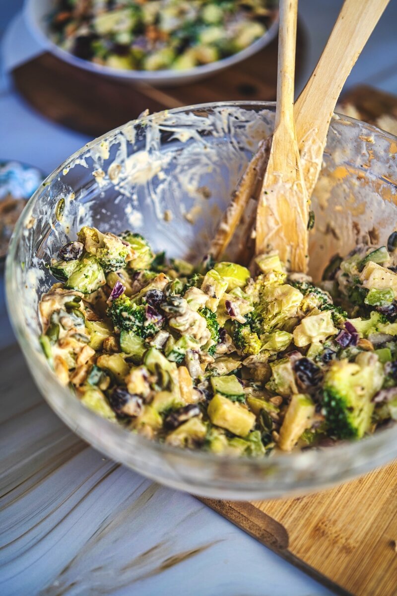 Creamy broccoli salad being mixed in a glass bowl with vegan dressing, crunchy vegetables, nuts, and seeds for an easy plant-based salad recipe