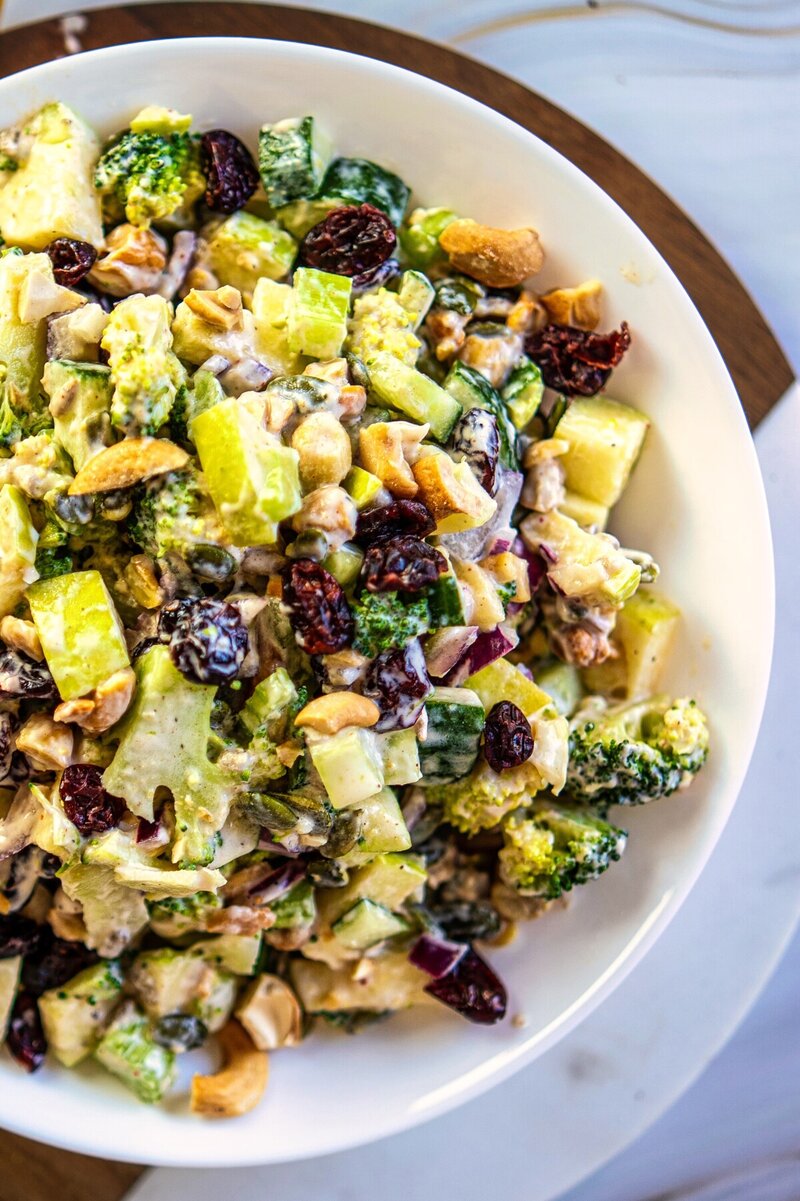 Creamy vegan broccoli salad with cucumber, green apple, dried cranberries, roasted cashews, sunflower seeds, and pumpkin seeds, served in a white bowl