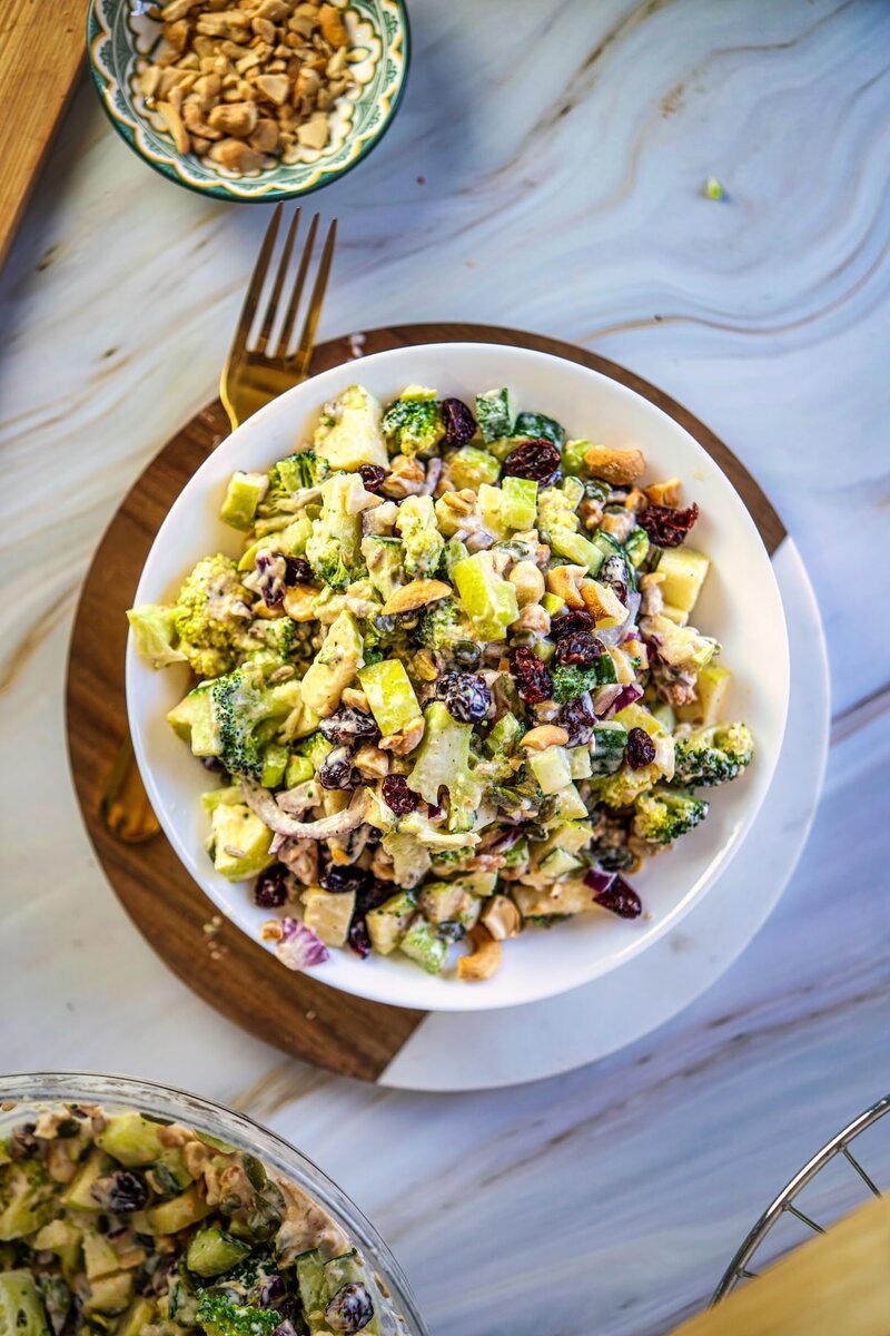 Overhead view of creamy broccoli salad in a bowl with green apple, cucumber, dried cranberries, roasted cashews, and seeds for a healthy vegan salad