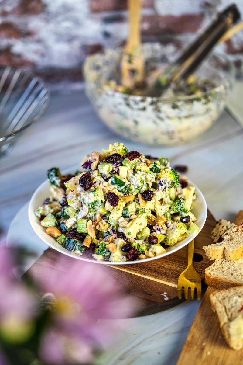 Homemade creamy broccoli salad on a wooden board with bread slices, made with blanched broccoli, crunchy vegetables, nuts, and vegan dressing