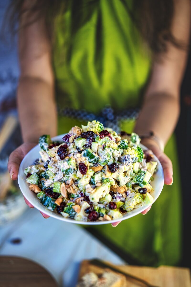 Vegan broccoli salad served on a plate, held by hands, featuring blanched broccoli, apple, cucumber, cranberries, and creamy dairy-free dressing