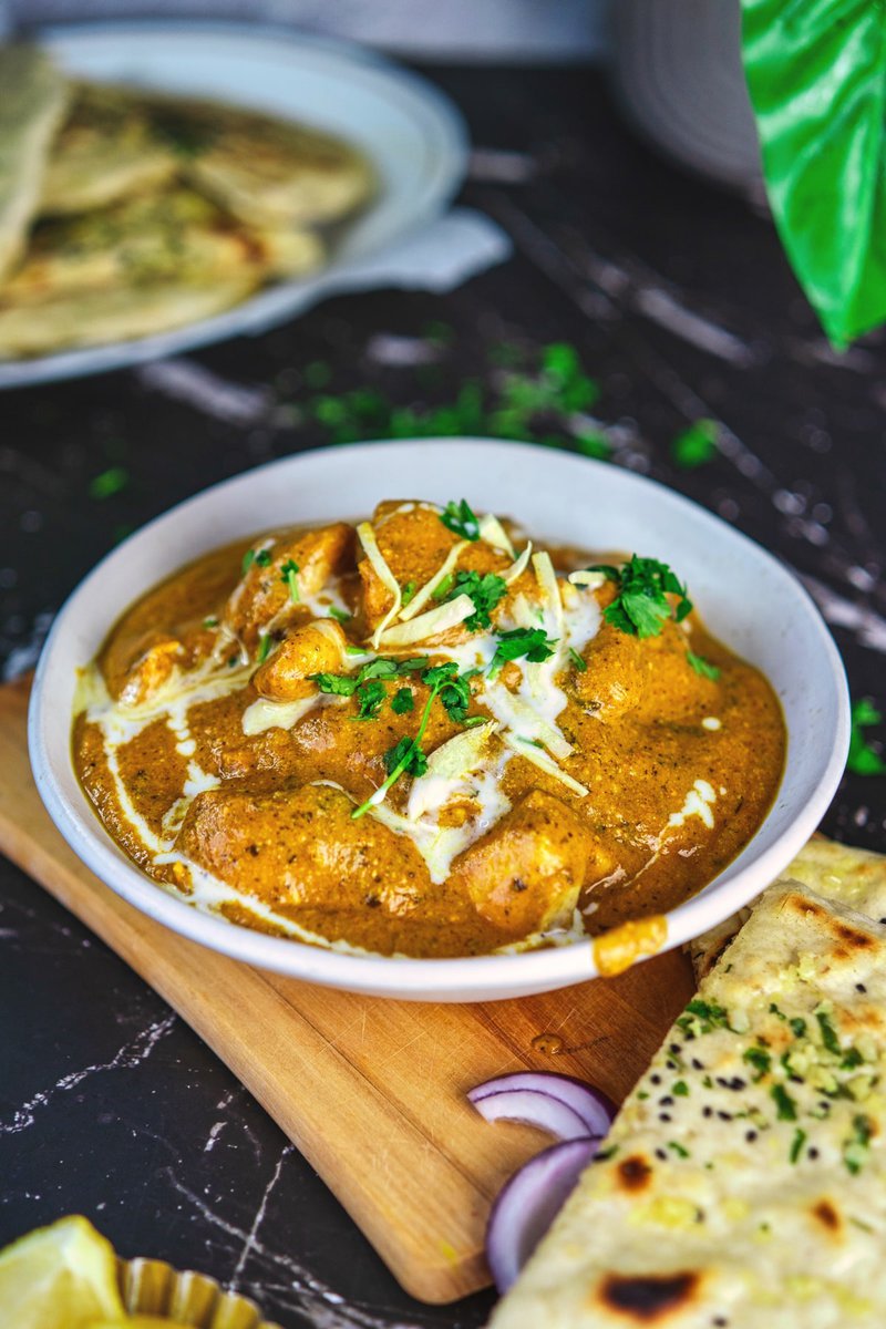 A bowl of creamy butter chicken garnished with cilantro, accompanied by garlic naan, on a dark background.