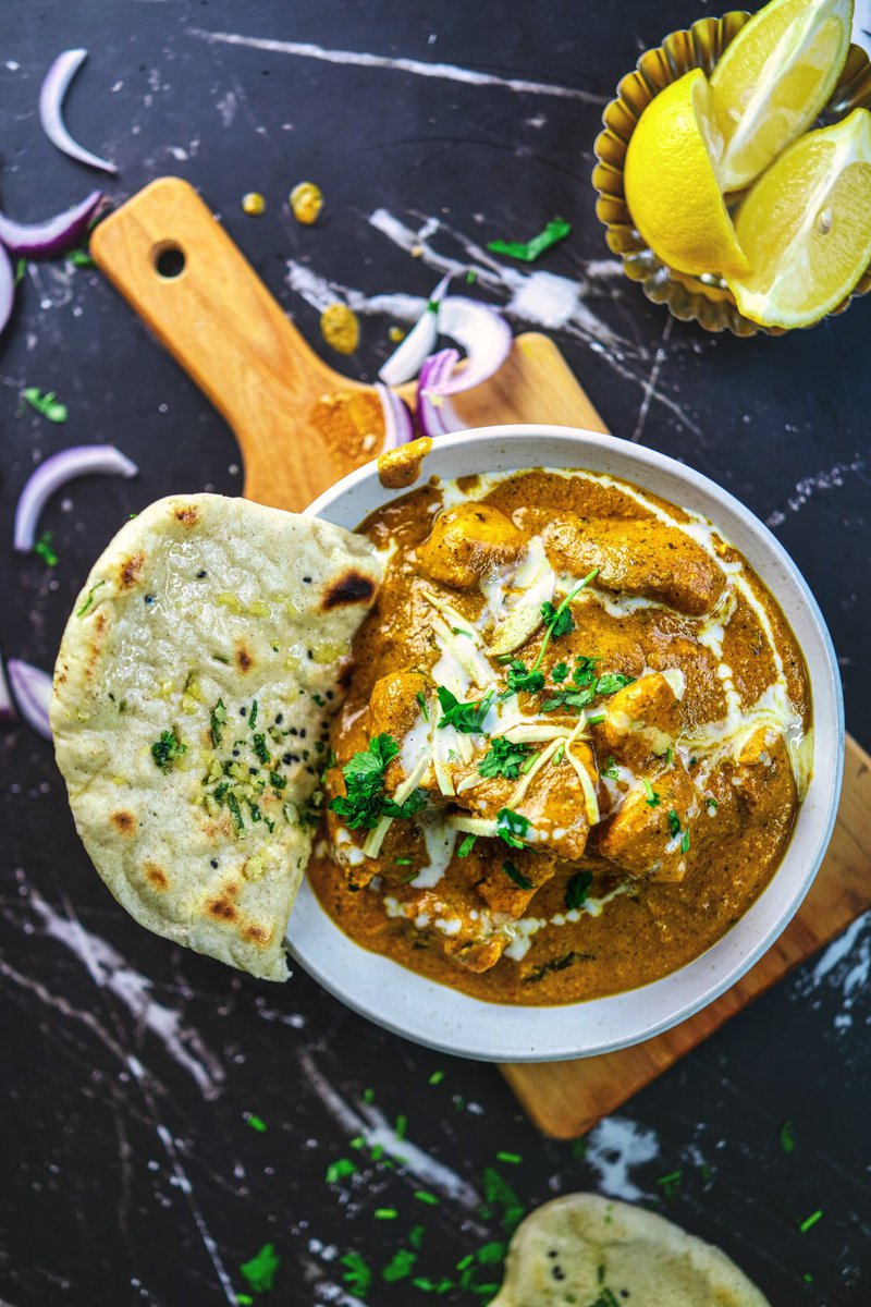 Butter chicken served in a white bowl with a side of garlic naan, lemon slices, and red onion on a wooden board.