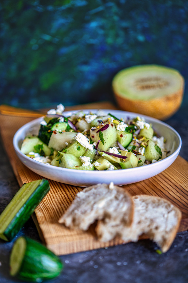 Elegant presentation of Cantaloupe melon salad in a bowl, garnished with feta cheese and pistachios, with bread and cucumber on the side.
