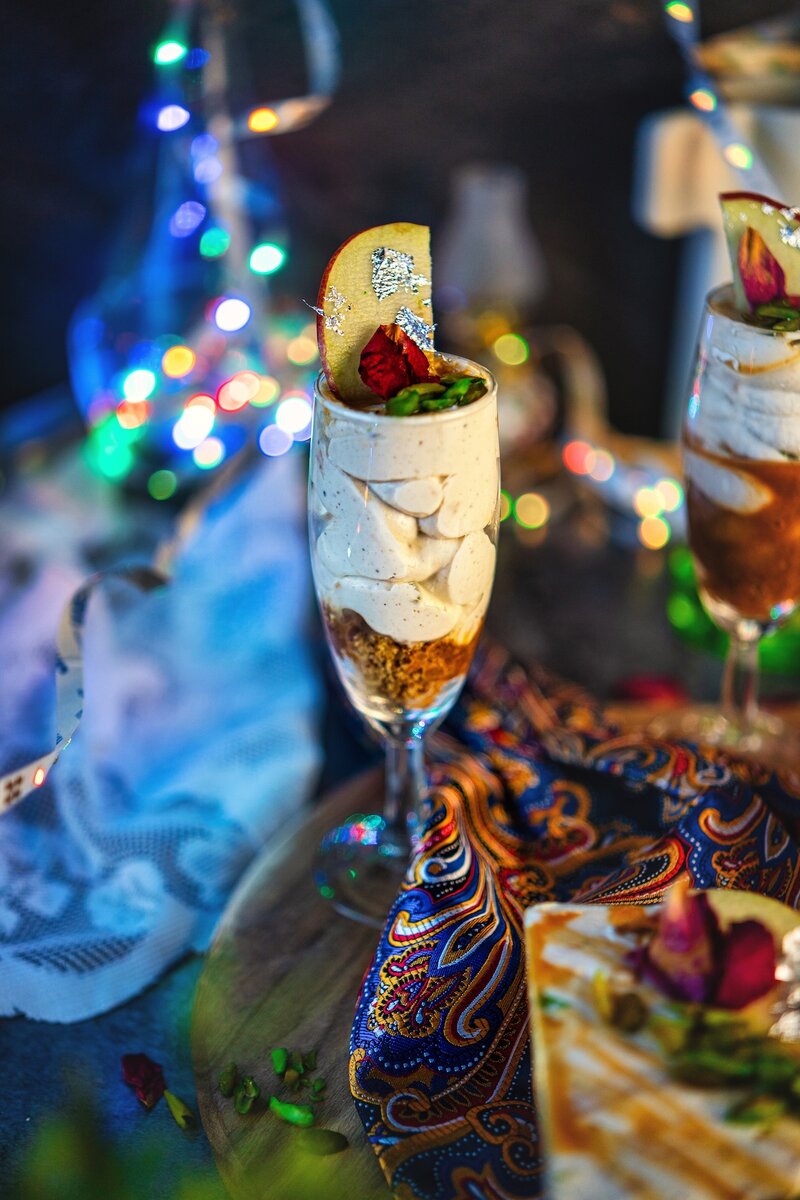 Caramel Apple Shrikhand served in a tall champagne glass with creamy Greek yogurt layers, cookie crumble base, caramel drizzle, pistachios, apple slice, and silver leaf.
