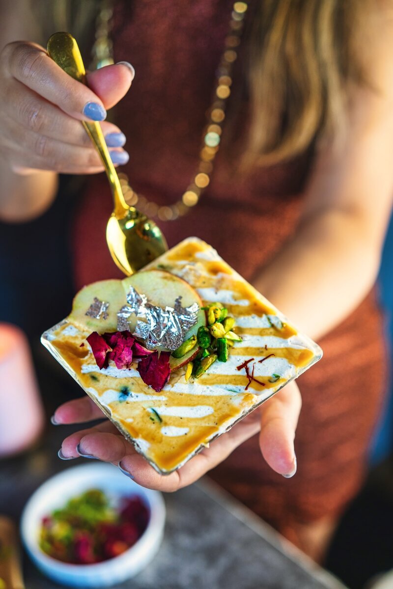 Hand holding Caramel Apple Shrikhand topped with apple slices, pistachios, silver leaf, and rose petals — a festive Indian fusion dessert.