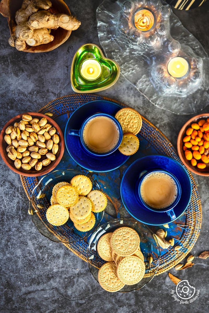 two cups of caramel chai and some cookies are on a tray next to a candle and a bowl of nuts with a candle in the background