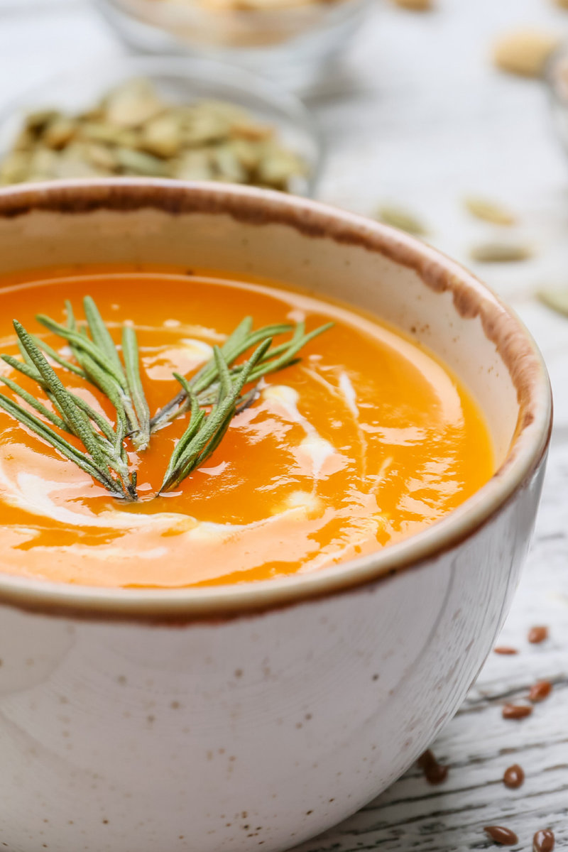 A close-up of creamy carrot gazpacho garnished with rosemary in a ceramic bowl, with a backdrop of scattered pumpkin seeds and a hint of fresh ingredients.