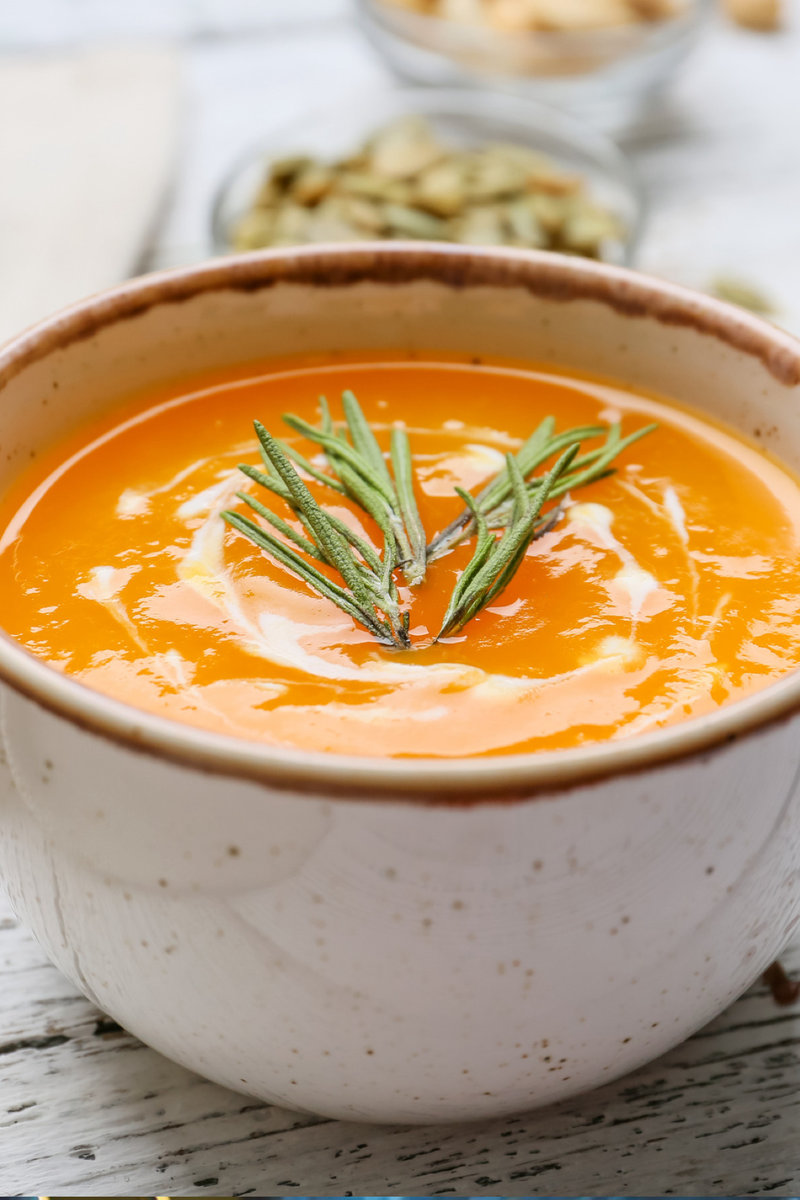 A vibrant carrot gazpacho served in a rustic bowl topped with a sprig of rosemary, set against a white wooden background with light natural tones.