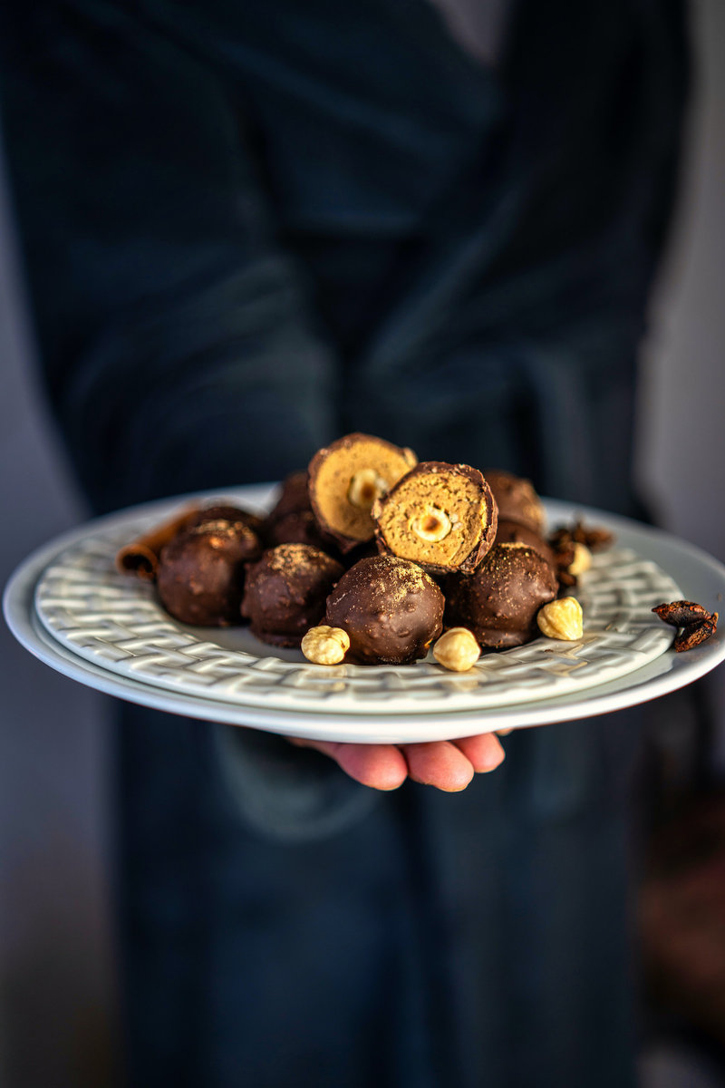 A hand holding a plate of chai truffles. The truffles are made with a dark chocolate shell and a chai-infused filling. They are garnished with crushed hazelnuts.