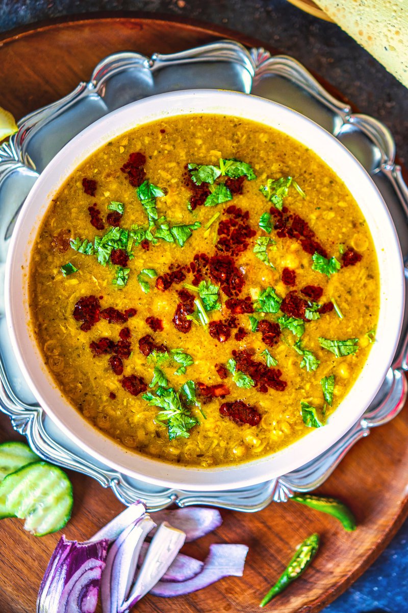 :A white bowl of chana dal garnished with fresh cilantro and a red chili tempering, placed on a silver plate with cucumber slices and onion pieces on a wooden tray.