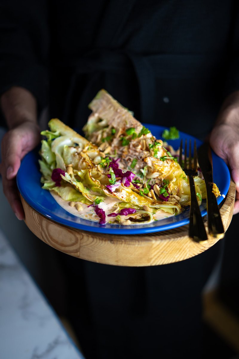 Serving charred cabbage wedges with pink sauce, chopped peanuts and fresh herbs on wooden board with blue plate