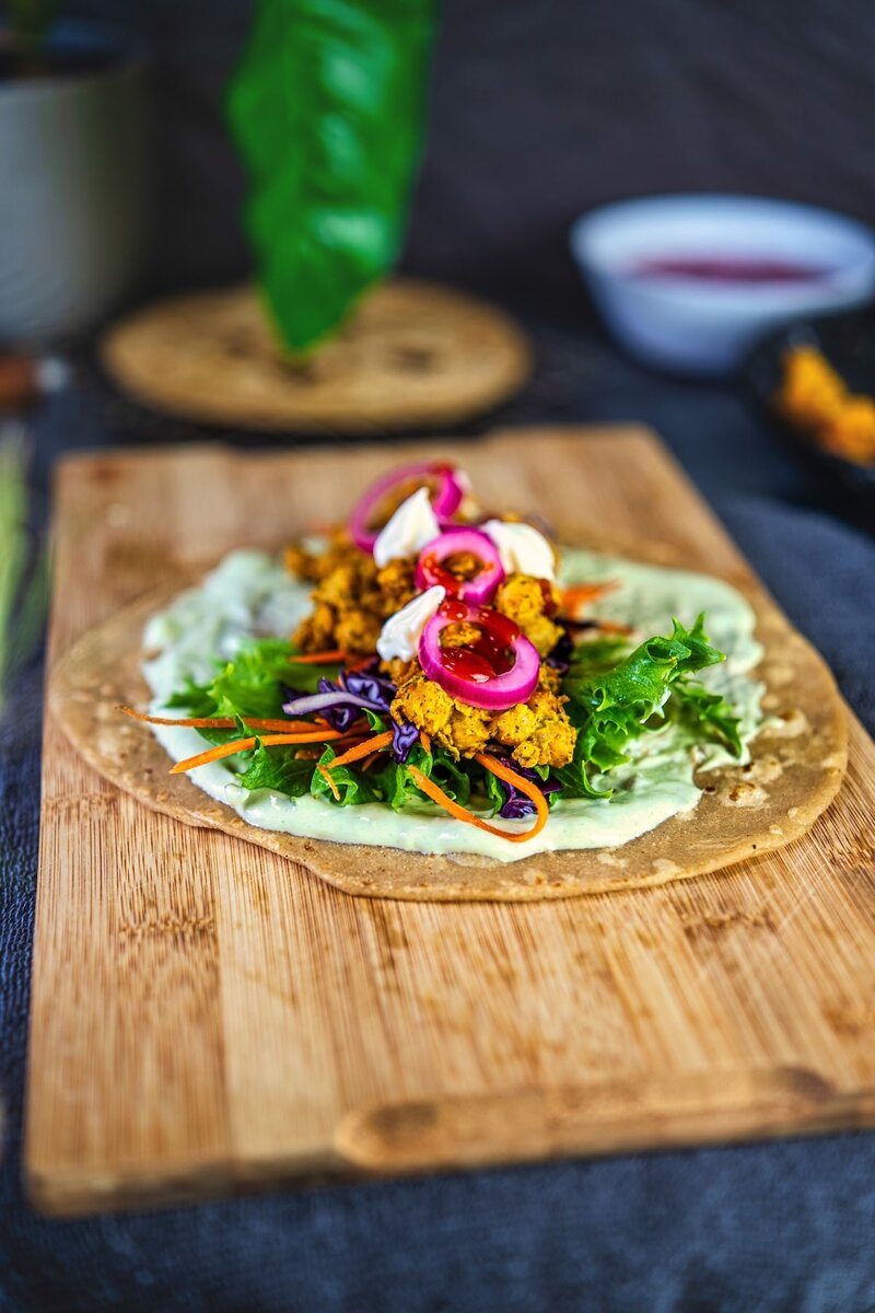 Chickpea tikka wrap on a wooden board, topped with fresh lettuce, carrots, purple cabbage, pickled onions, vegan mayonnaise, and green yogurt sauce