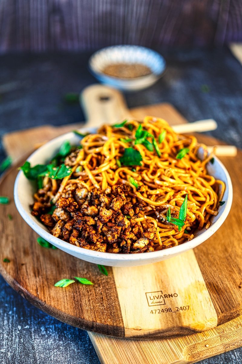 Bowl of Chili Garlic Noodles with crispy tofu, sesame seeds, and spring onions on a wooden board