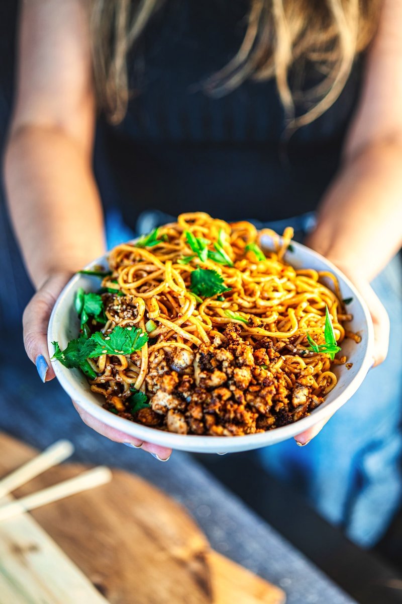 Hands holding a bowl of Chili Garlic Noodles with crispy tofu, spring onions, cilantro, and sesame seeds