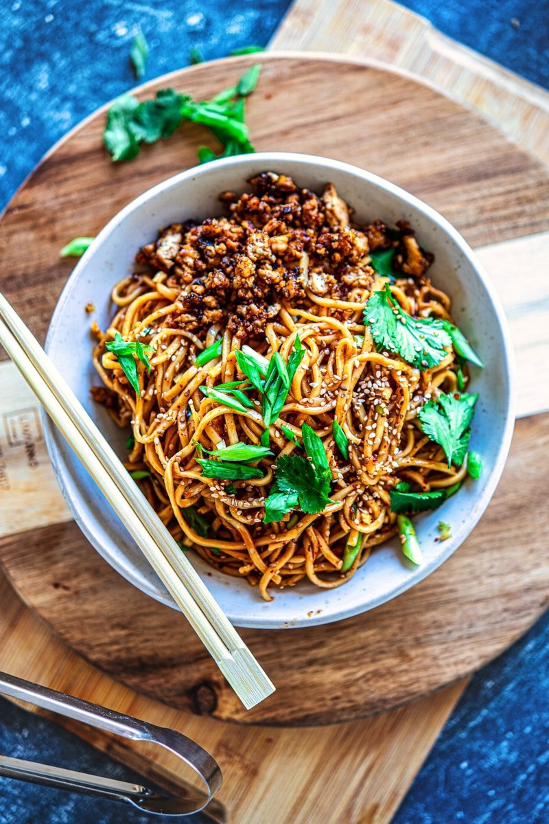 Chili Garlic Noodles with crispy tofu, sesame seeds, spring onions, and chopsticks on a wooden serving board