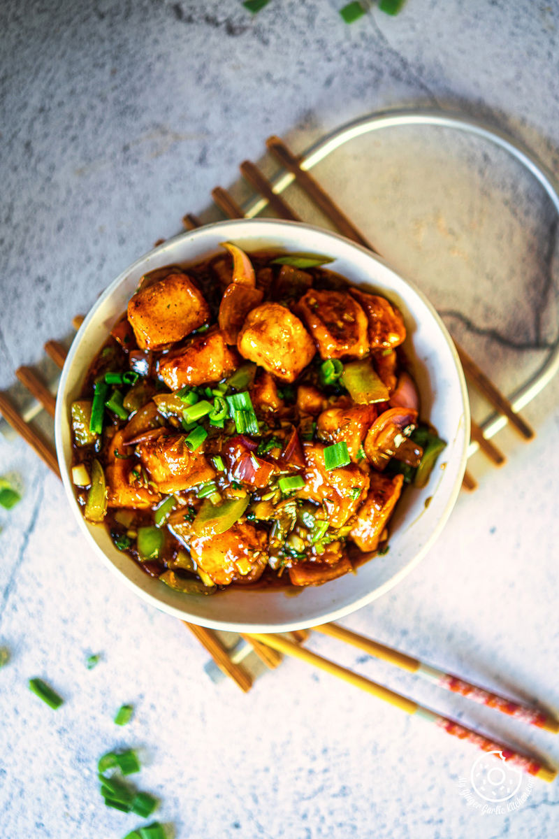 overhead photo of a bowl of chilli paneer with chopsticks and a plate of food